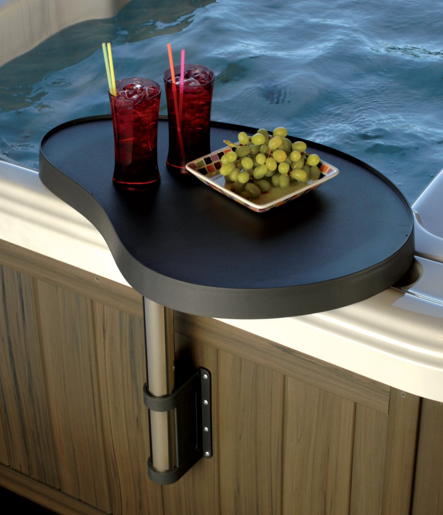 A black SpaCaddy with a sturdy reinforced polymer tray on an aluminum post, mounted on a wooden spa wall. There are two red beverage cups and a dish of green grapes on the tray.